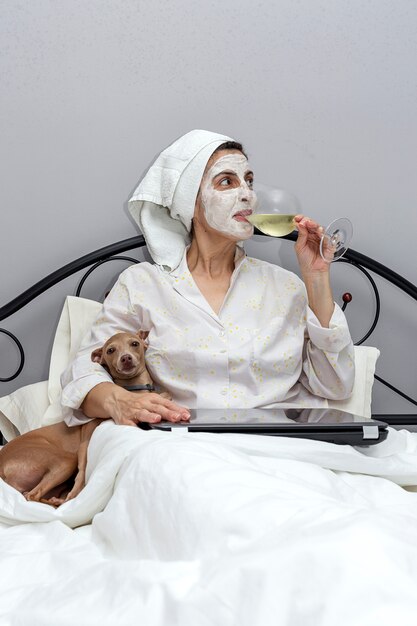 Mujer cuidando su rostro con una mascarilla en la cama, con perro, bebiendo una copa de vino