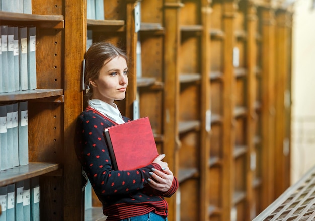 Mujer curiosa de pie cerca de los estantes de la biblioteca con libro rojo