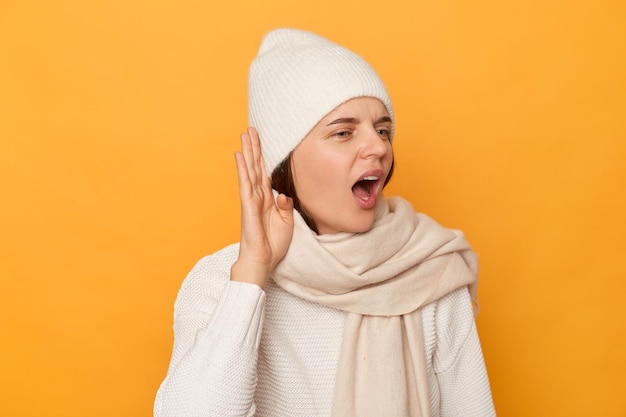 Mujer curiosa con ropa abrigada mantiene la mano cerca de la oreja tratando de escuchar rumores tiene una expresión intrigada tratando de escuchar una conversación posando aislada sobre un fondo amarillo