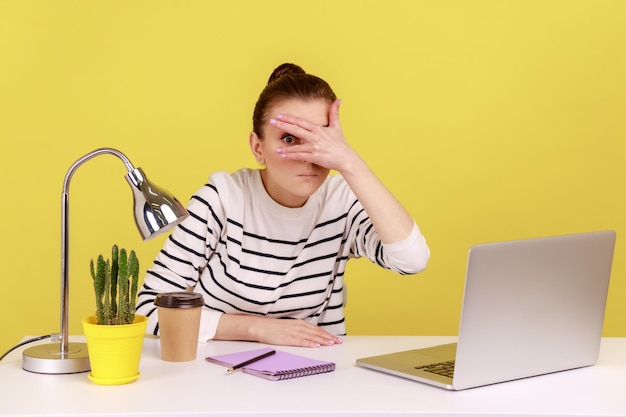 Mujer curiosa sentada en el lugar de trabajo con una computadora portátil y espiando mirando a través de los dedos con interés