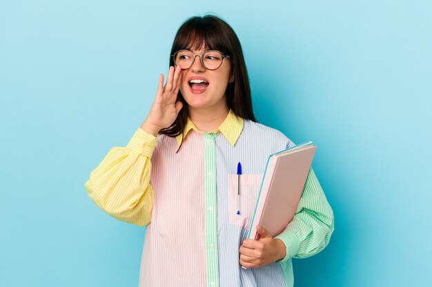 Mujer con curvas joven estudiante sosteniendo libros aislados sobre fondo azul gritando y sosteniendo la palma cerca de la boca abierta.