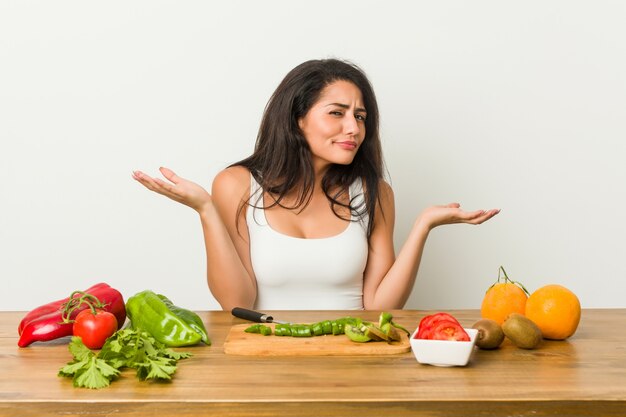 Mujer con curvas joven que prepara una comida sana que duda y que se encoge de hombros en gesto inquisitivo.