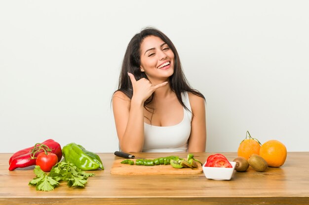 Mujer con curvas joven que prepara una comida sana que muestra un gesto de llamada de teléfono móvil con los dedos.