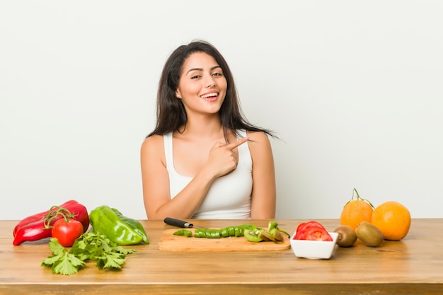 Mujer con curvas joven que prepara una comida sana que sonríe y que señala a un lado, mostrando algo en el espacio en blanco.