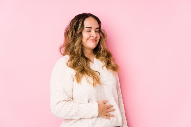La mujer con curvas joven que presenta en una pared rosada aislada toca la panza, sonríe suavemente, comiendo y concepto de la satisfacción.