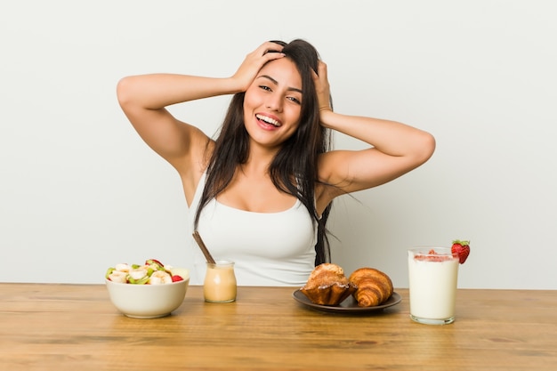 La mujer con curvas joven que toma un desayuno ríe alegremente manteniendo las manos en la cabeza. Concepto de felicidad