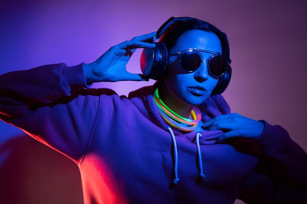 Una mujer cyberpunk con capucha y gafas de sol baila contra la pared con palos de neón y auriculares