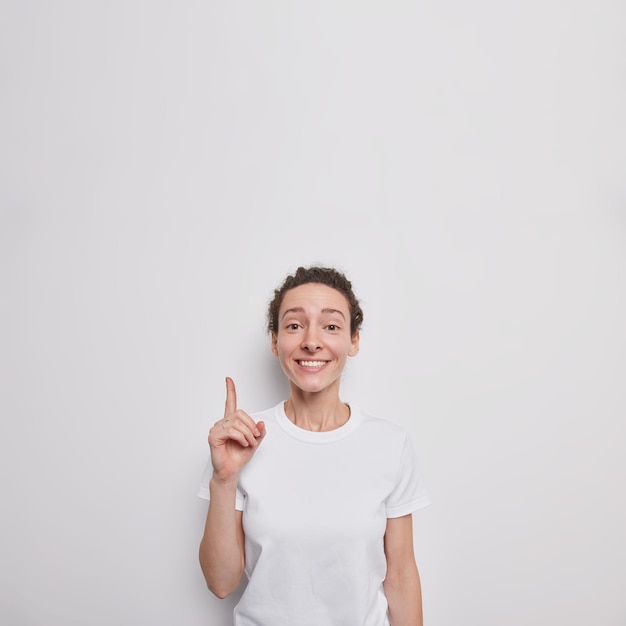 La mujer da recomendaciones señala el dedo índice arriba en el espacio de la copia en blanco sonríe agradablemente muestra el espacio de la copia viste una camiseta informal aislada sobre una pared blanca