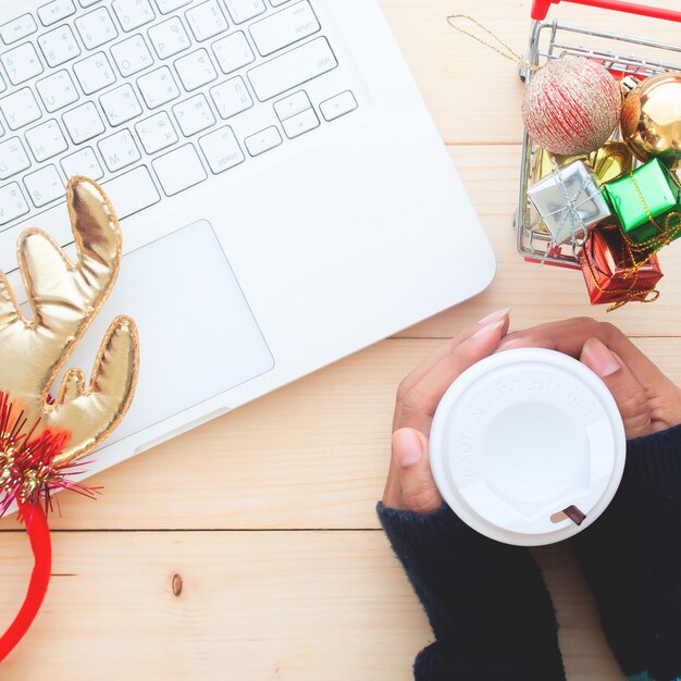 La mujer da sostener la taza de café en la tabla con el ordenador portátil, concepto de las compras de la Navidad