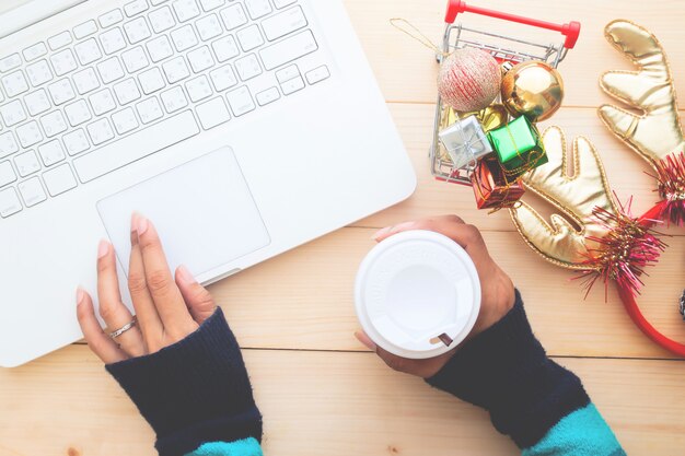 La mujer da sostener la taza de café en la tabla con el ordenador portátil, concepto de las compras de la Navidad