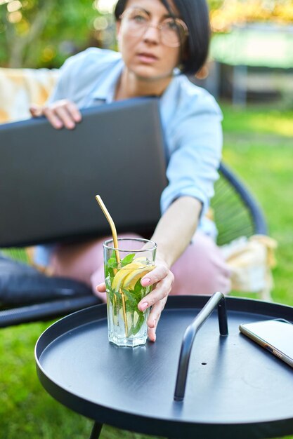 La mujer deja la computadora portátil y toma limonada sentada en una silla en el patio trasero