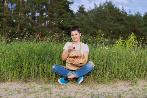 Mujer en un delantal con cesta de fresas frescas, fondo de paisaje natural. Alimentos orgánicos saludables, agricultura y pequeñas empresas, jardinería y pasatiempos.