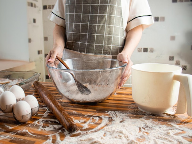 Foto mujer en un delantal en la cocina prepara la masa de harina huevos de harina y un cuenco transparente de vidrio cerca de cocinar en casa