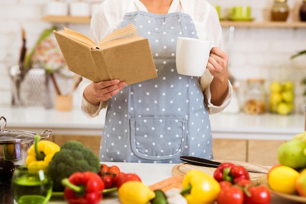 Una mujer con un delantal lee un libro de cocina mientras sostiene una taza en su cocina