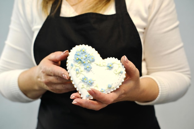 Una mujer con un delantal negro sostiene un pan de jengibre en forma de corazón con flores pintadas en sus manos Foto horizontal