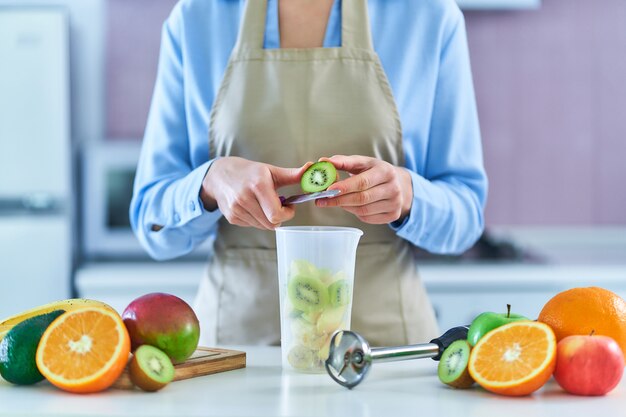 Mujer en delantal preparando un batido de frutas frescas en su casa en la cocina con batidora de mano