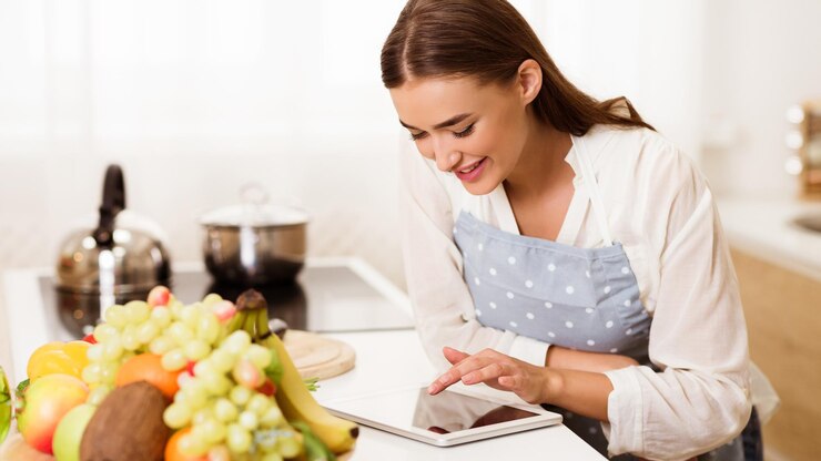 Foto una mujer con un delantal se para en su cocina y usa una tableta para encontrar una receta