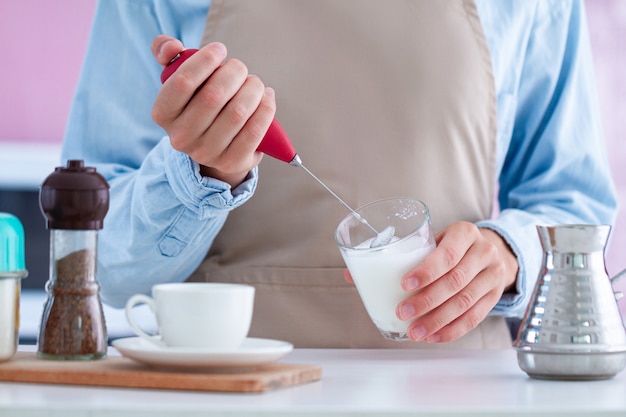 Mujer en delantal con un vaporizador de leche para hacer café en la cocina en casa