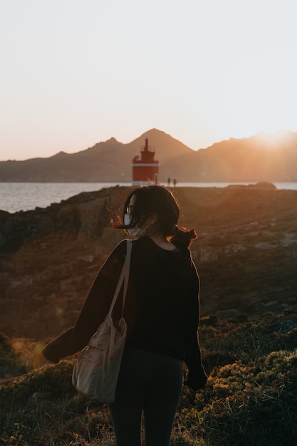 Mujer delante de un faro rojo durante una colorida puesta de sol con una máscara