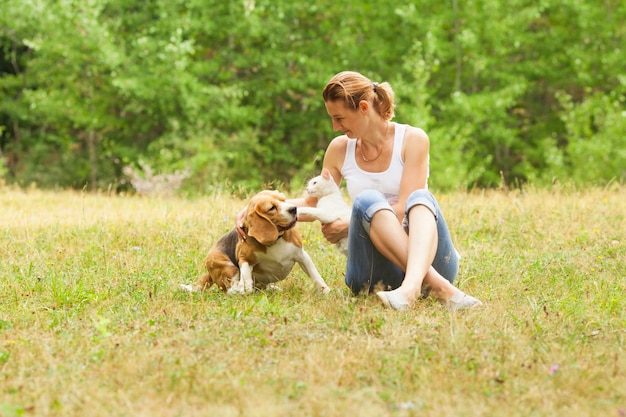 Mujer delgada atractiva joven sentada en la hierba jugando con su gato y su perro. Los animales domésticos y su dueño se divierten al aire libre en el prado de verano con un bosque verde al fondo