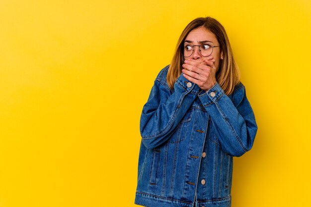 Mujer delgada caucásica joven aislada en la pared amarilla pensativa mirando a un espacio de la copia que cubre la boca con la mano.