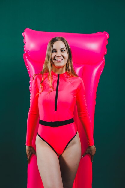 Una mujer delgada y feliz con un traje de baño rosa brillante sostiene y posa con un colchón inflable rosa sobre un fondo verde aislado Colchón de aire rosa Colchón de piscina de verano