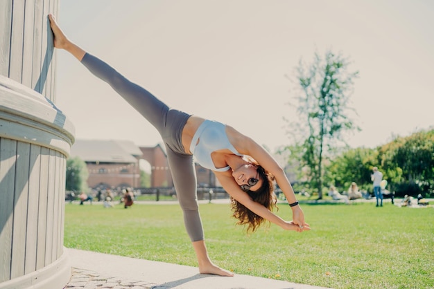 Mujer delgada flexible que hace ejercicios de estiramiento al aire libre, se mantiene en buena forma física vestida con un top corto y leggings se para en una pierna se inclina con los brazos levantados se calienta antes del entrenamiento cardiovascular