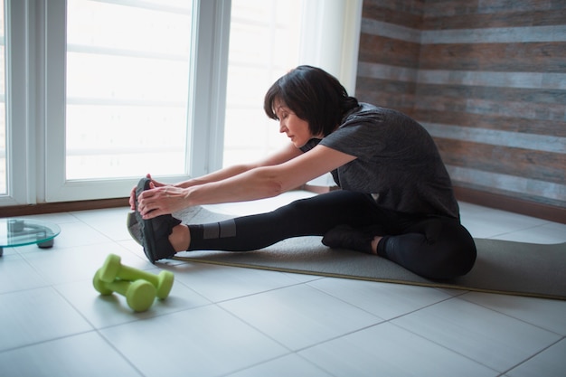Mujer delgada en forma adulta tiene entrenamiento en casa. Foto de modelo senior sentada en una estera de yoga y estirando hacia adelante hasta los dedos de los pies. Cuida el bienestar del cuerpo.