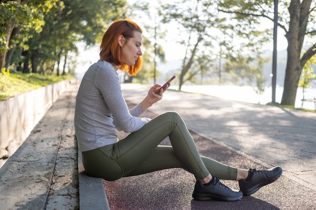 Mujer delgada de jengibre en ropa deportiva con teléfono móvil en el parque. Salud, bienestar, estilo de vida fitness.