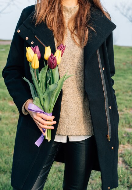 Mujer delgada joven en un abrigo que se coloca en un prado verde y que sostiene un ramo de tulipanes disponible