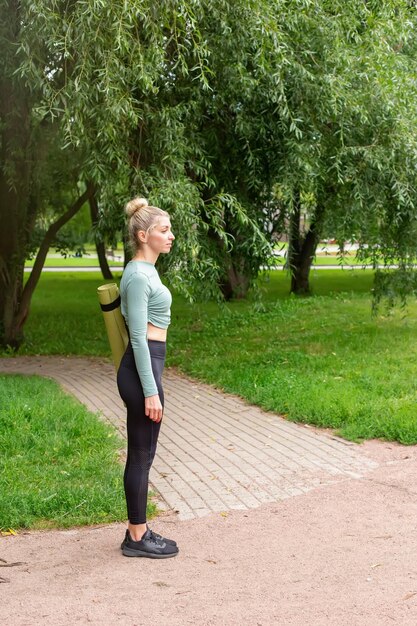 Una mujer delgada con ropa deportiva de pie en verano en el parque con una alfombra de gimnasio verde para deportes