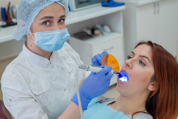 Mujer dentista empastes dentales fijos y secos con luz Hermosa mujer dentista poniendo un empaste en un diente con caries de una paciente pelirroja
