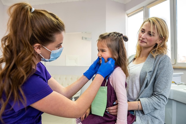 Mujer dentista examinando los dientes de una niña en la clínica dental Madre apoyando a su pequeña hija en el consultorio del dentista