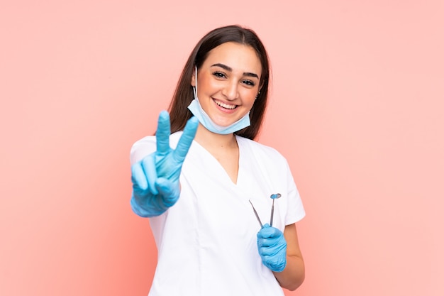 Mujer dentista con herramientas aisladas en pared rosa sonriendo y mostrando el signo de la victoria