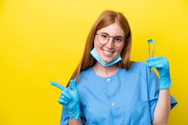 Mujer dentista joven pelirroja aislada sobre fondo amarillo señalando con el dedo al lado