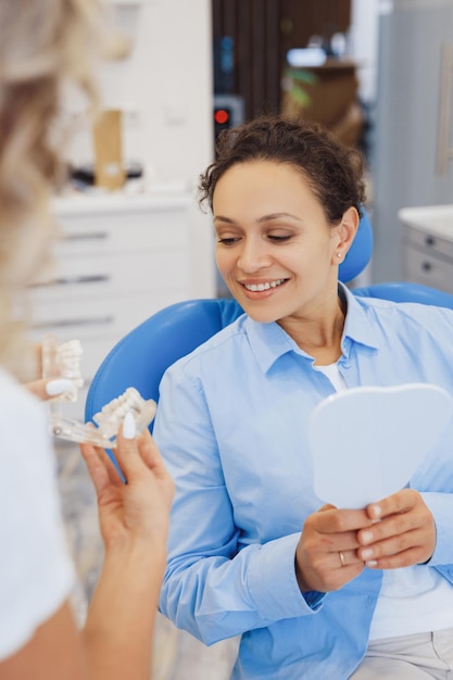 Mujer dentista mostrando modelo de mandíbulas en las manos Cliente alegre discutiendo procedimientos médicos en el consultorio dental