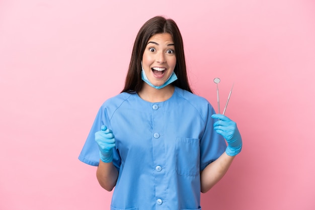 Mujer dentista sosteniendo herramientas aisladas sobre fondo rosa celebrando una victoria en la posición ganadora