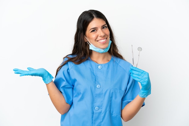 Mujer dentista sosteniendo herramientas sobre fondo blanco aislado con expresión facial conmocionada