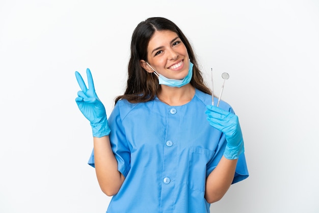 Mujer dentista sosteniendo herramientas sobre fondo blanco aislado sonriendo y mostrando el signo de la victoria