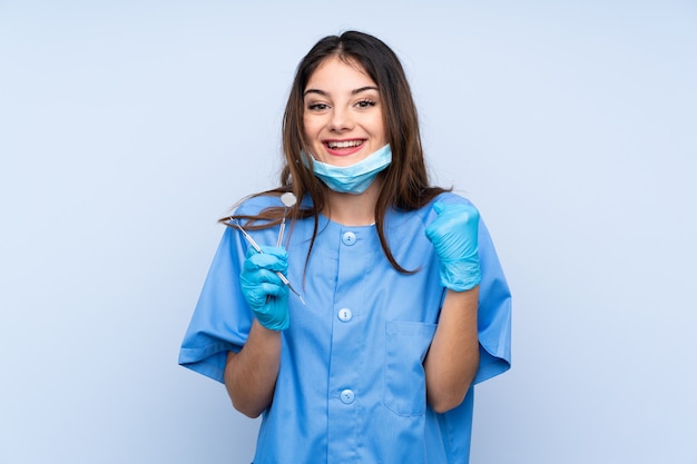 Mujer dentista sosteniendo herramientas sobre pared azul aislado celebrando una victoria