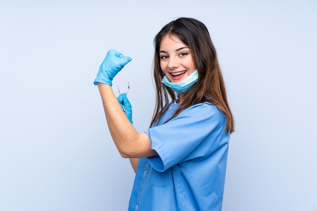 Mujer dentista sosteniendo herramientas sobre pared azul haciendo un gesto fuerte