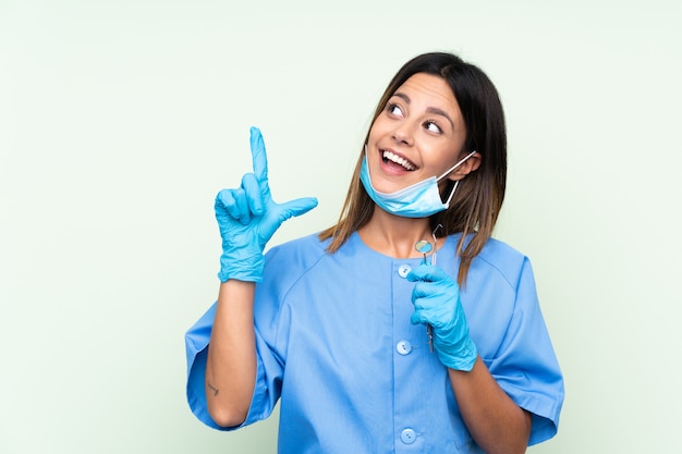 Mujer dentista sosteniendo herramientas sobre pared verde aislado señalando con el dedo índice una gran idea