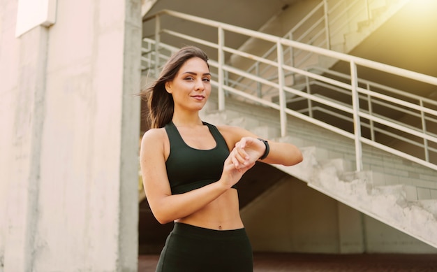 Mujer de deporte atractivo en ropa deportiva utiliza reloj inteligente al aire libre en el entorno urbano