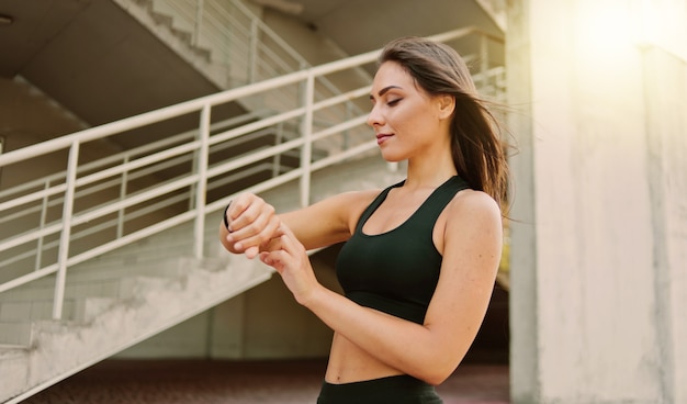 Mujer de deporte atractivo en ropa deportiva utiliza reloj inteligente al aire libre en el entorno urbano