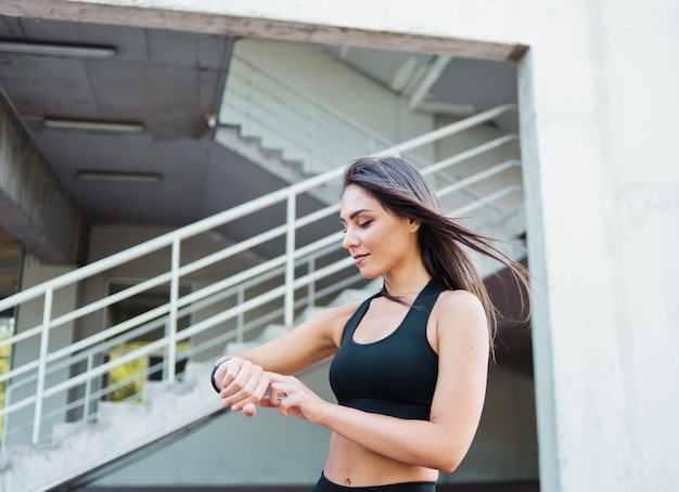 Mujer de deporte atractivo en ropa deportiva utiliza reloj inteligente al aire libre en el entorno urbano