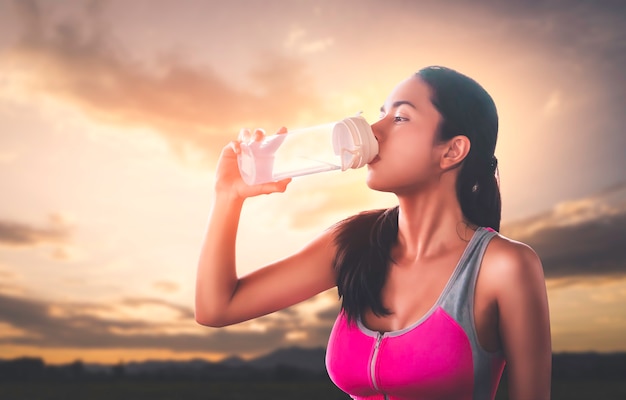 La mujer del deporte está bebiendo agua dulce sobre un fondo de cielo de nubes doradas.