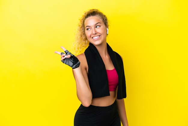 Mujer de deporte con toalla aislado sobre fondo amarillo sonriendo y mostrando el signo de la victoria