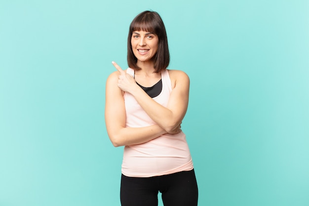 Foto mujer deportista sonriendo alegremente, sintiéndose feliz y apuntando hacia un lado y hacia arriba, mostrando el objeto en el espacio de la copia