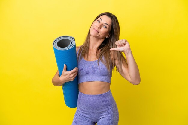 Mujer deportista yendo a clases de yoga mientras sostiene una alfombra aislada de fondo no amarillo orgullosa y satisfecha