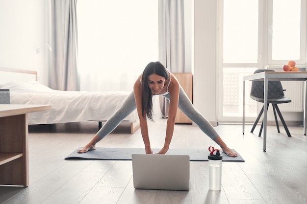 Mujer deportiva. Alegre morena de pelo largo de pie sobre la alfombra y mirando directamente a la computadora portátil, mientras estira su cuerpo